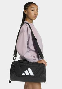Bolsa de viaje negra con logo de Adidas blanco, cuenta con correa ajustable, bolsillos con cremallera y un material duradero con textura suave.
