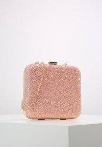 Pochette à paillettes roses de forme carrée, avec des accents métalliques et une bandoulière en or. La surface présente une finition texturée et scintillante.