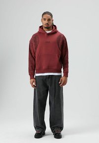 Sweat à capuche bordeaux avec un motif latéral, une poche avant et un logo. Porté avec un jean foncé ample et des chaussures noires avec des lacets bien visibles.