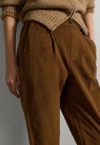 Pantaloni in corduroy marroni con una finitura texture, dotati di tasche laterali e una vestibilità ampia. L'elastico in vita e le pieghe sottili aumentano il comfort.