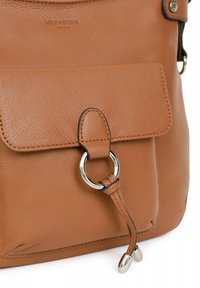 Borsa a tracolla in pelle beige con tasca frontale, anello in metallo argentato e un nodo decorativo in pelle con accenti metallici. Dettaglio della superficie testurizzata visibile.