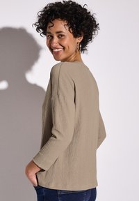 Beige texturovaný top s dlouhým rukávem a kulatým výstřihem, volný střih a jemným bočním rozparkem, kombinovaný s tmavými džíny. Model se usmívá.