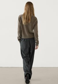 Maglione marrone lavorato a maglia con orlo a costine, abbinato a pantaloni grigi scuri a righe sottili. Capelli lunghi e lisci; in piedi visto da dietro.