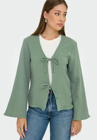 Cardigan verde texturizado con escote en V y cierre frontal atado. Mangas largas con un diseño fluido, llevado sobre una camiseta blanca y jeans.
