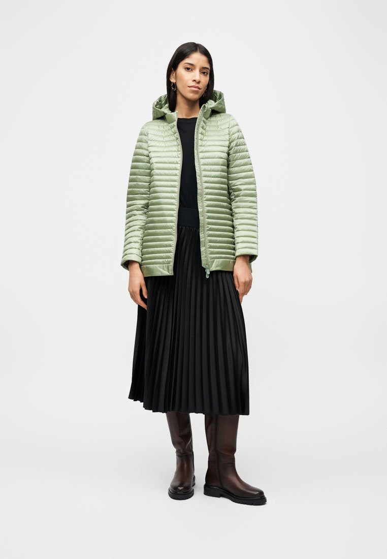 Femme portant une doudoune vert clair sur un haut noir et une jupe midi plissée, associée à des bottes marron montantes, debout devant un fond blanc.