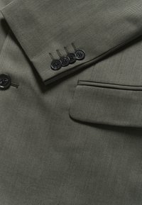 Atsitiktinis žalia mažo dydžio kostiumo švarkas su tekstūruotu audiniu, turintis penkis juodus mygtukus rankogaliuose, elegantišką apykaklę ir kišenę su apvadu.