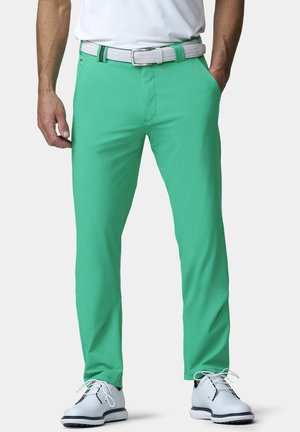 Mann trägt grüne Hose mit weißem geflochtenem Gürtel, weißes Hemd und weiße elegante Schuhe, Hände locker an den Seiten.
