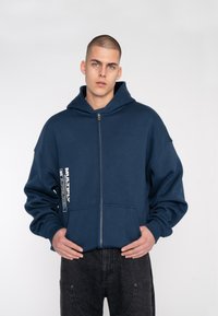 Haut en jersey bleu marine avec zip, fabriqué en tissu doux, doté d'une grande capuche, de poches avant et d'un détail d'étiquette imprimé sur le bras gauche.