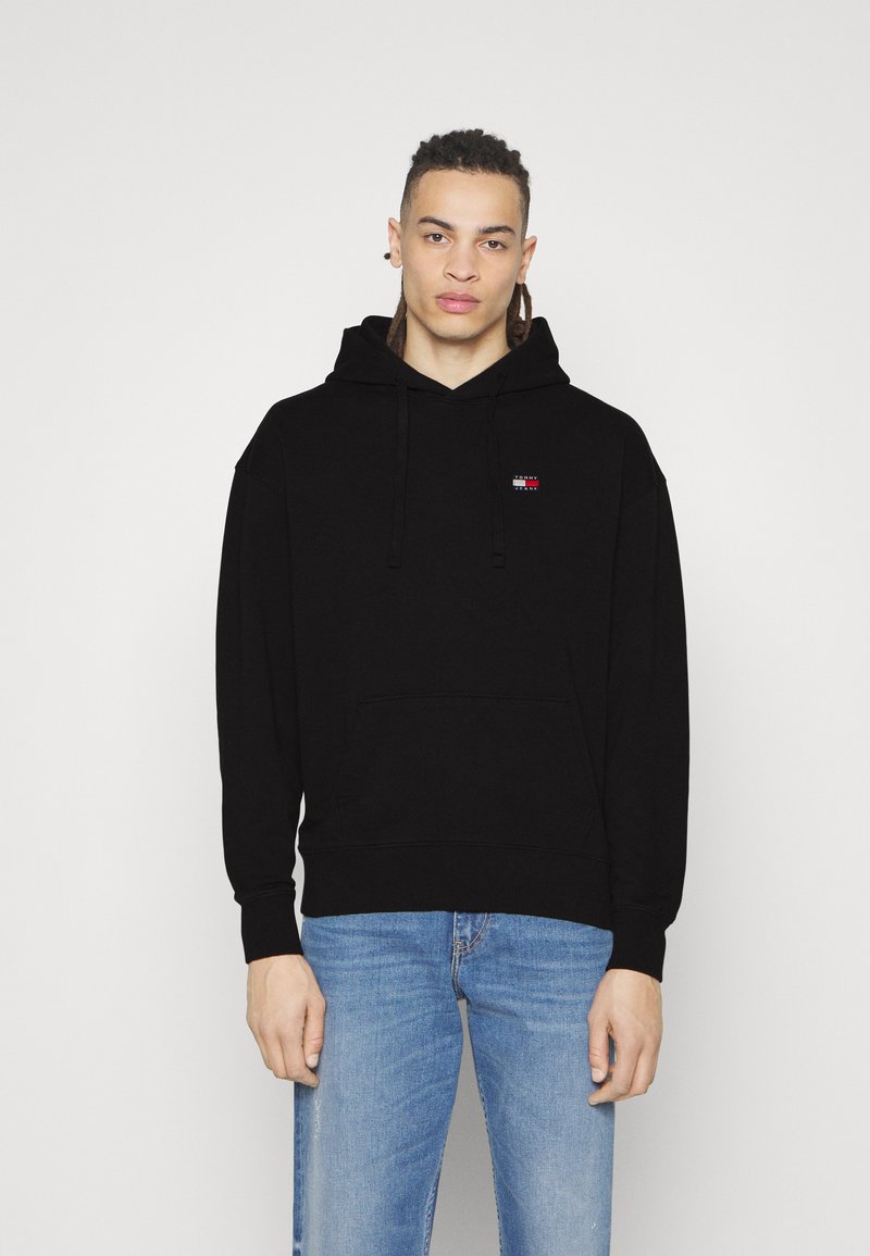 Schwarzer Hoodie aus Baumwolle mit einer Vordertasche und Kapuze mit Kordelzug. Enthält ein kleines mehrfarbiges Logo auf der Brust. Getragen mit blauen Jeans.