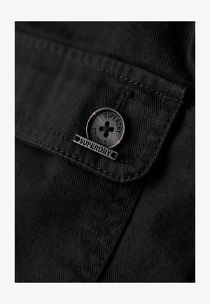 Black fabric featuring a close-up of a button with "Superdry" branding and a flap. Textured surface with contrasting stitching detail.