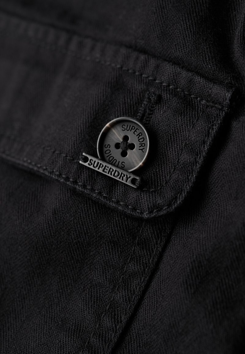 Tela negra con un primer plano de un botón con la marca "Superdry" y una solapa. Superficie texturizada con detalles de costura en contraste.
