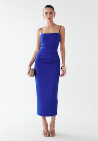 Mėlyna kūną apgaubiančio stiliaus midi ilgio suknelė su plonais petnešėliais, tekstūruotos medžiagos ir surinktais detalėmis. Derinama su auksinėmis dirželiais puoštomis sandalais ir smėlio spalvos delninuke.