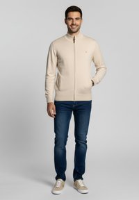 Suéter beige con cremallera y cuello alto, puños y dobladillo acanalados. Combinado con vaqueros azul oscuro y zapatillas de color claro.