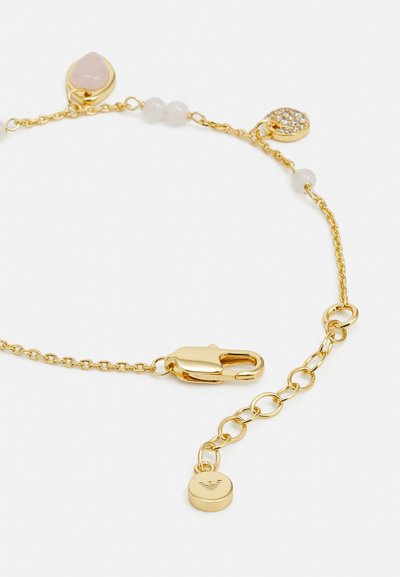 Gold chain bracelet featuring a lobster clasp, rose quartz charm, white beads, and a textured circular charm with logo detail.