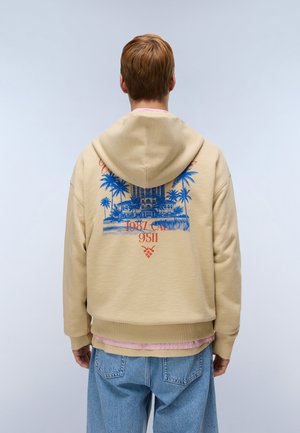 Person with short hair wears beige hoodie featuring blue palm trees and building graphic with red text on back, paired with light blue jeans.