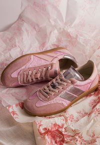 Paire de baskets roses en daim et satin avec semelles en caoutchouc marron et accents métalliques, reposant sur un papier froissé à motif floral.
