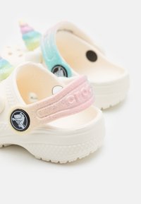 Weiße Kinderclogs mit bunten Akzenten in Form von Einhornhörnern. Die Riemen sind pastellblau und rosa mit Glitzer und zeigen ein rundes Logo-Detail.