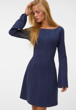 Femme aux longs cheveux blonds portant une robe bleu marine ajustée avec des manches longues légèrement évasées, touchant ses cheveux d'une main.