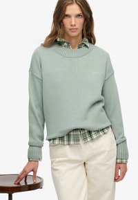 Mujer con un suéter de punto verde claro sobre una camisa de cuadros y pantalones crema, reposando la mano izquierda sobre un taburete de madera redondo.