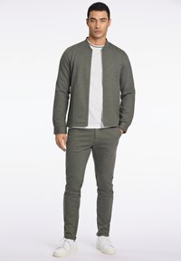 Chaqueta de punto verde oscuro con cuello alzado y cremallera, combinada con pantalones a juego. Camiseta blanca de cuello redondo por debajo y zapatillas blancas.