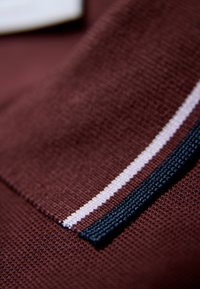 Bordowa tkanina teksturowana z paskowym akcentem w białym i ciemnoniebieskim kolorze. Zbliżenie na szczegóły ściegów.