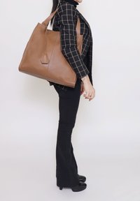 Bruine leren shopper met een gestructureerde afwerking, zeshoekige metalen details en stevige schouderbanden, afgebeeld tegen een effen achtergrond.