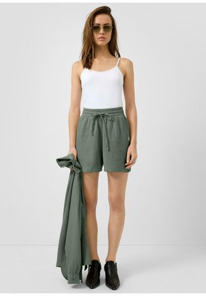 Młoda kobieta ubrana w biały top na ramiączkach, oliwkowe szorty, trzymająca pasującą koszulę z długim rękawem i czarne skórzane botki z ćwiekami, stoi w pomieszczeniu.