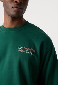 Sweatshirt vert avec col côtelé, arborant un texte brodé "Société des Brew du Matin Paresseux" en orange et blanc. Texture lisse.
