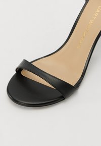 Fekete bőr magassarkú szandál minimalista dizájnnal, vékony pánttal a lábujjak felett és sima, bézs talpbéléssel.
