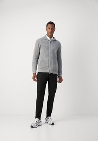 Siva rebrasta zip-up kapuca z dvema sprednjima žepoma, v kombinaciji s črnimi hlačami in belimi supergami z sivimi akcenti. Oplazni ozadje.