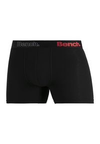 Black boxer shorts made of soft fabric, featuring a mid-rise waistband with "Bench." logo in black and red. Plain design with no patterns.