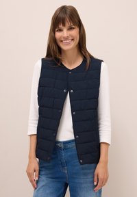 Gilet smanicato trapuntato blu navy con scollatura rotonda, bottoni a pressione e cuciture sottili, indossato sopra una maglietta bianca a maniche lunghe, abbinato a un jeans.