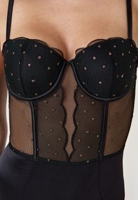 Schwarzes Korsettoberteil mit transparenten Mesh-Einsätzen, gewelltem Spitzenrand und kleinen rosé-goldenen Punkten. Verfügt über verstellbare Träger und ein glattes, figurbetontes Design.
