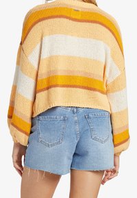 Stickad tröja med gula, orange och vita ränder, med en beskuren nederkant och ballongärmar, som är ihop med ljusblå denimshorts.