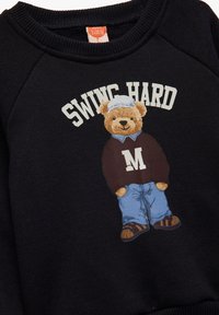 Sudadera negra con un gráfico de un oso de peluche con un suéter burdeos con la letra "M" y jeans azules. El texto dice "SWING HARD". Tejido suave y texturizado.