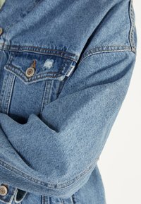 Jeansjacka i ljusblått tyg, med knappstängning fram, två bröstfickor, slitna detaljer och synlig sömnad.