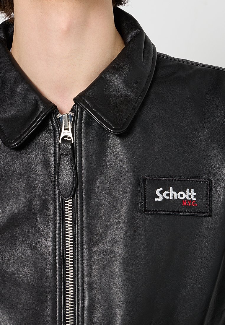 Gros plan sur une veste en cuir noire avec une fermeture éclair argentée et un patch rectangulaire du logo Schott N.Y.C. sur la poitrine, portée par une personne.