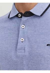 Ljust blå strukturad polo-shirt med svart randsydd krage och tre vita knappar knäppta vid halsen.