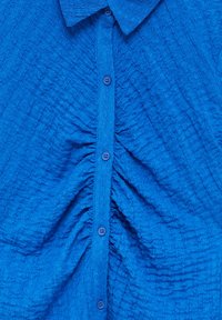 Chemise boutonnée en tissu bleu texturé, avec un col, des boutons sur le devant et un détail froncé au centre.