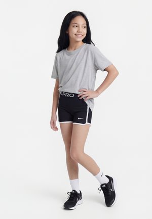 Djevojka stoji s rukom na bokovima, nosi sivu majicu, crne Nike Pro kratke hlače, bijele čarape i crne Nike tenisice na bijeloj pozadini.