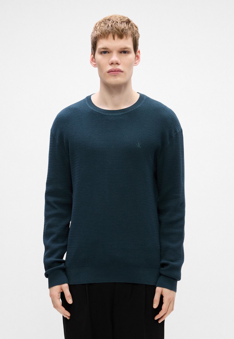 Knit sweater in dark teal with a textured pattern, crew neckline, and long sleeves. Ribbed cuffs and hem, minimal branding visible.