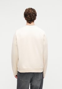 Beige sweatshirt med rund hals og ribbestrikkede mansjetter, som viser en jevn tekstur. Brukt med mørkegrå bukser, sett bakfra.