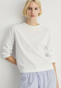 White long-sleeve T-shirt made of cotton, featuring a round neckline and rolled cuffs, paired with blue and white striped pajama pants.