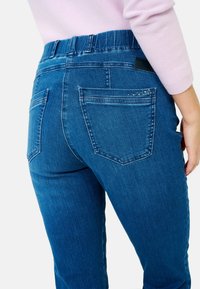 Jean en denim bleu avec une taille élastique, comportant deux poches arrière et des strass décoratifs près de la poche droite.