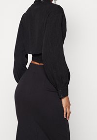 Chemisier noir court en tissu texturé avec des manches bouffantes, associé à une jupe noire ajustée. Détail de boutons discret sur les poignets de la chemise.