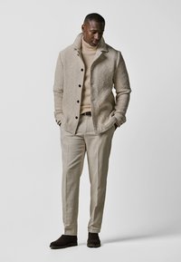 Beige Strickjacke mit Kragen und schwarzen Knöpfen, getragen über einem hellbeigen Rollkragenpullover, kombiniert mit hellbeigen Hosen und braunen Wildlederstiefeln.