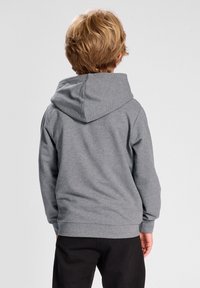 Felpa grigia con un grande cappuccio arrotondato, texture morbida e polsini a coste. Vista da dietro, abbinata a pantaloni neri.