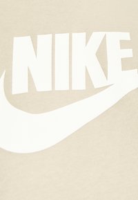 Světlé béžové bavlněné tričko s velkým bílým nápisem "NIKE" a pod ním designem swoosh, zdůrazňující minimalistickou značku a hladkou texturu.