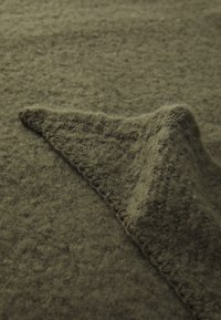 Couverture en laine de couleur vert olive atténué, présentant une surface texturée et un bord cousu à la main, avec un coin plié révélant des fibres douces.