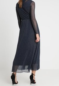 Robe longue navy en tissu texturé, manches longues transparentes, taille froncée et léger éclat métallique. Associée à des sandales noires à talons hauts.